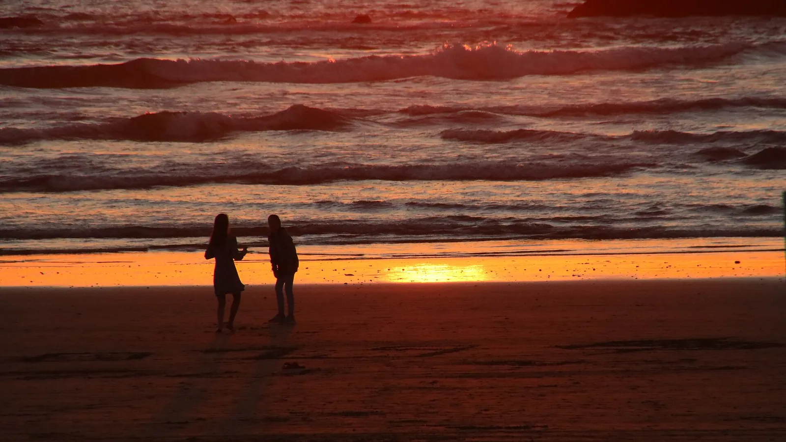 Two people silhouetted on Agadir beach as the sun sets over Atlantic waves