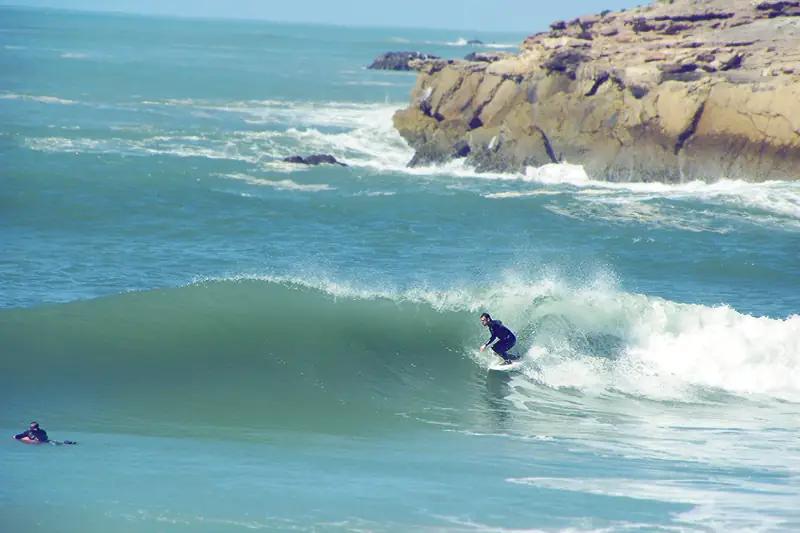 A surfer riding a green Atlantic wave beside a rocky point near Tamraght